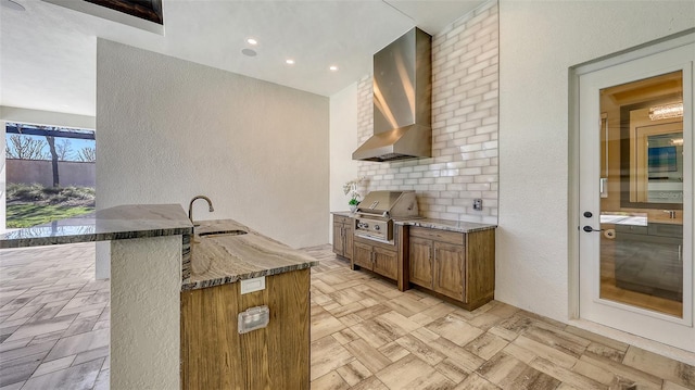 kitchen with brown cabinets, a sink, tasteful backsplash, dark stone counters, and wall chimney exhaust hood