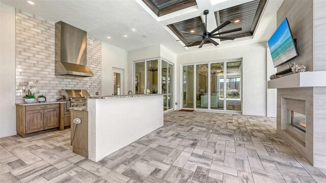 kitchen featuring brown cabinetry, a ceiling fan, coffered ceiling, a glass covered fireplace, and wall chimney range hood