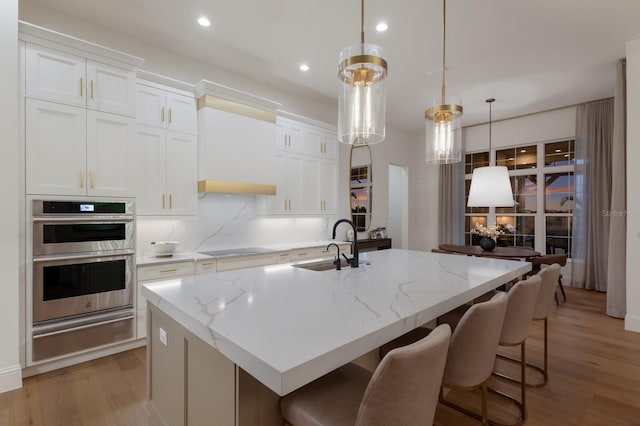 kitchen with a warming drawer, a kitchen island with sink, a sink, stainless steel double oven, and light wood finished floors