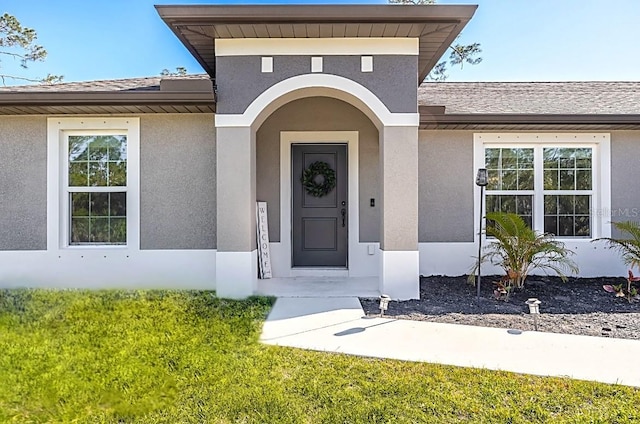 entrance to property featuring stucco siding, a shingled roof, and a yard