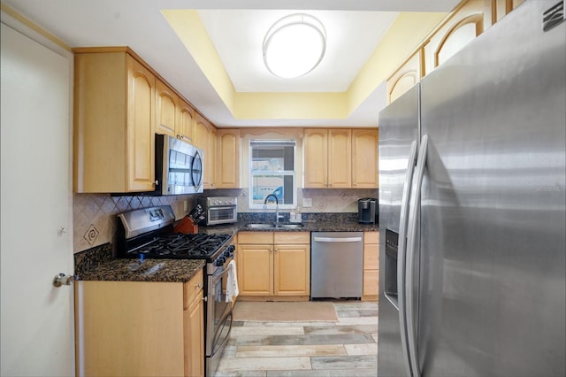 kitchen with light brown cabinets, a sink, stainless steel appliances, a raised ceiling, and tasteful backsplash