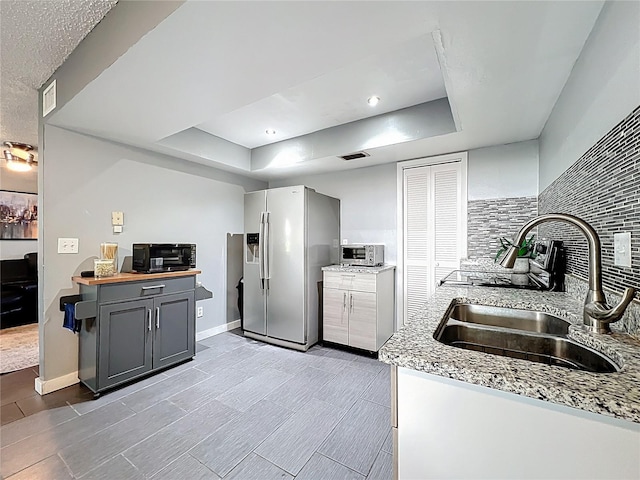 kitchen with backsplash, gray cabinetry, stainless steel fridge, a raised ceiling, and a sink