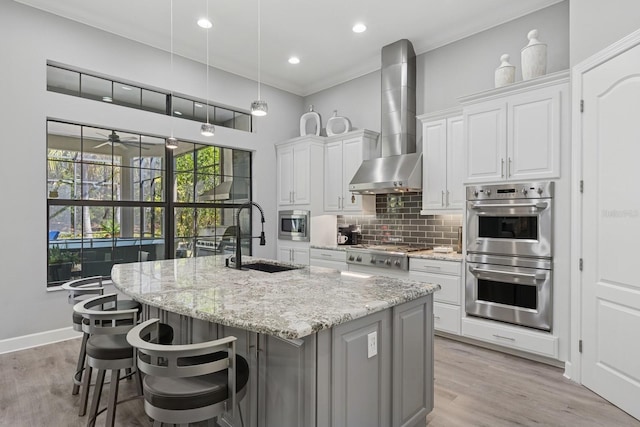 kitchen featuring a kitchen island with sink, a sink, appliances with stainless steel finishes, wall chimney exhaust hood, and decorative backsplash