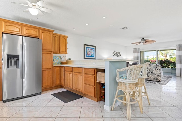 kitchen with a kitchen bar, light countertops, a peninsula, stainless steel refrigerator with ice dispenser, and a ceiling fan