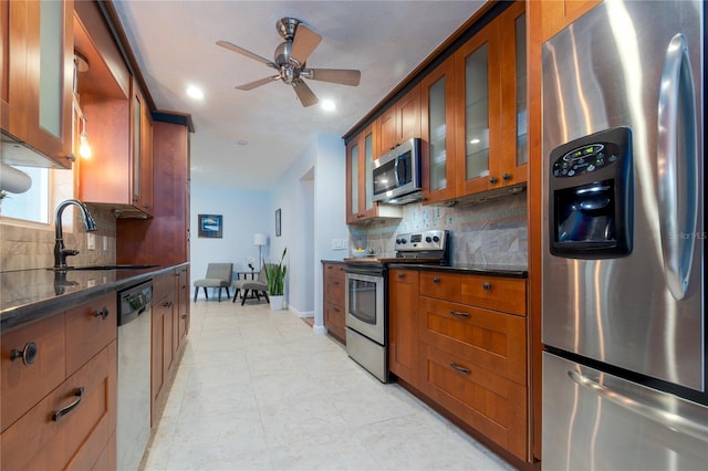 kitchen with a ceiling fan, brown cabinetry, a sink, stainless steel appliances, and glass insert cabinets