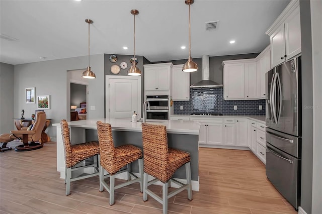 kitchen featuring tasteful backsplash, wall chimney range hood, light wood-type flooring, light countertops, and appliances with stainless steel finishes