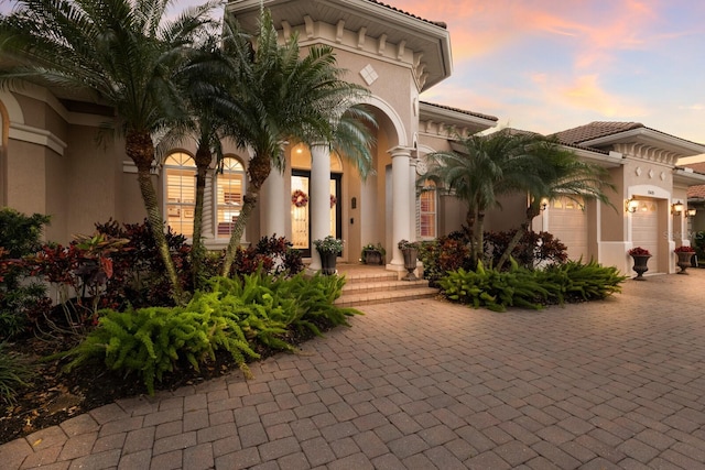 exterior entry at dusk with a tiled roof, stucco siding, an attached garage, and decorative driveway