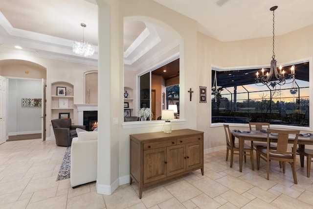 dining room with built in features, arched walkways, a lit fireplace, a raised ceiling, and a chandelier