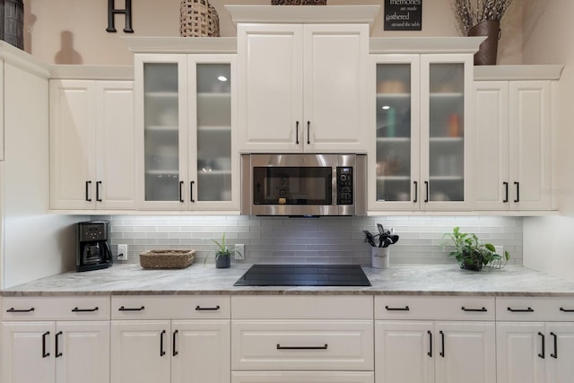 kitchen featuring stainless steel microwave, backsplash, white cabinetry, glass insert cabinets, and black electric stovetop