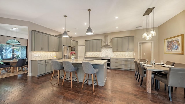 kitchen featuring wall chimney range hood, dark wood finished floors, stainless steel appliances, gray cabinetry, and a kitchen breakfast bar