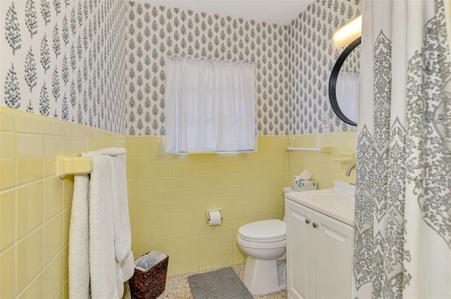 bathroom featuring wallpapered walls, tile walls, a wainscoted wall, toilet, and vanity