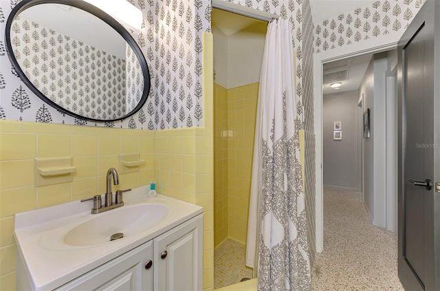 full bath featuring vanity, wallpapered walls, a tile shower, wainscoting, and tile walls