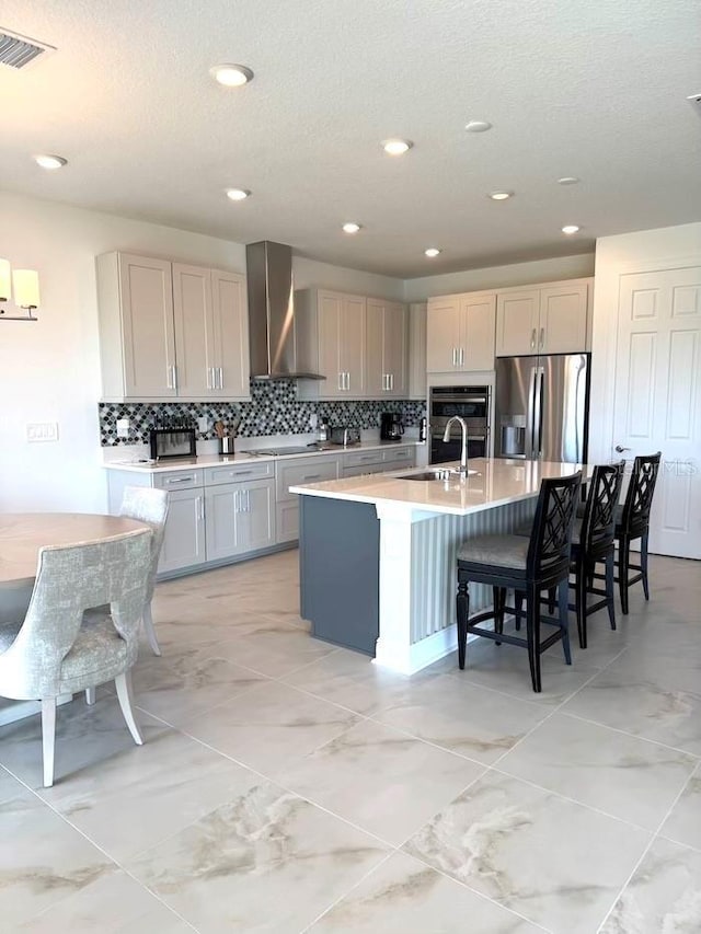 kitchen featuring a center island with sink, a breakfast bar, light countertops, appliances with stainless steel finishes, and wall chimney exhaust hood