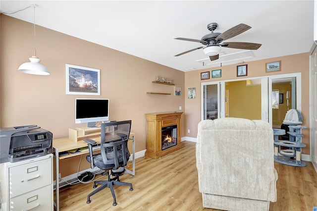 office featuring a glass covered fireplace, light wood-style flooring, baseboards, and ceiling fan