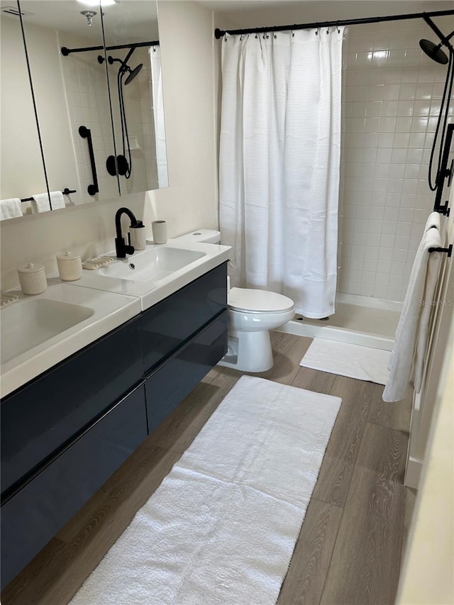 full bathroom featuring toilet, double vanity, a tile shower, wood finished floors, and a sink