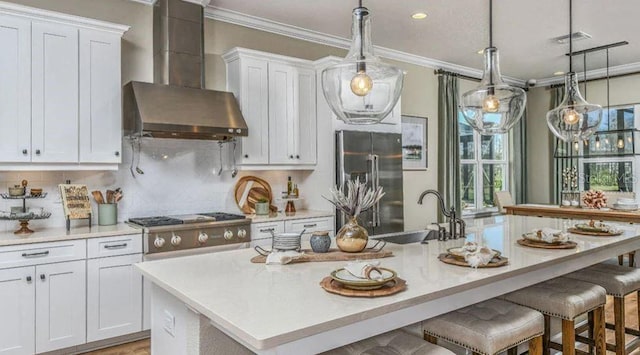 kitchen featuring an island with sink, ornamental molding, a sink, appliances with stainless steel finishes, and wall chimney exhaust hood