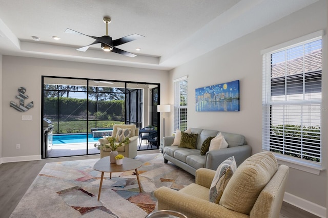 living room featuring baseboards, a ceiling fan, a tray ceiling, and wood finished floors