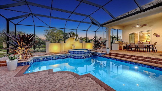 pool at dusk featuring glass enclosure, a patio, an outdoor kitchen, a pool with connected hot tub, and ceiling fan