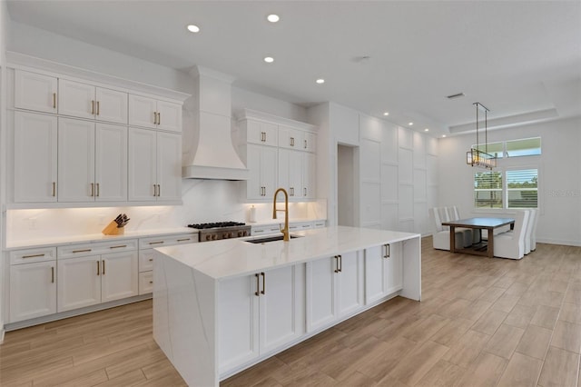 kitchen with light wood finished floors, a sink, premium range hood, white cabinets, and a kitchen island with sink