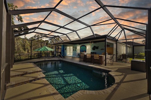 pool at dusk featuring glass enclosure, a patio, an outdoor pool, and outdoor dry bar