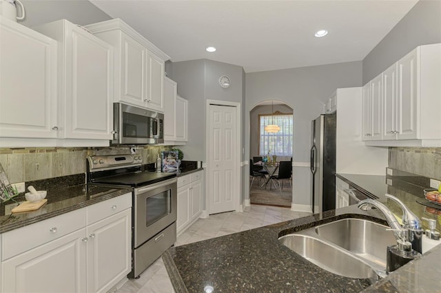 kitchen with a sink, white cabinetry, stainless steel appliances, arched walkways, and dark stone counters