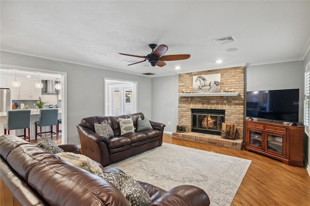 living room with visible vents, light wood finished floors, ceiling fan, crown molding, and a brick fireplace