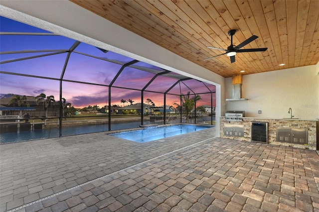 pool at dusk with a patio, grilling area, exterior kitchen, a sink, and glass enclosure