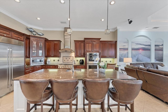 kitchen featuring wall chimney range hood, a breakfast bar, a sink, decorative backsplash, and built in appliances