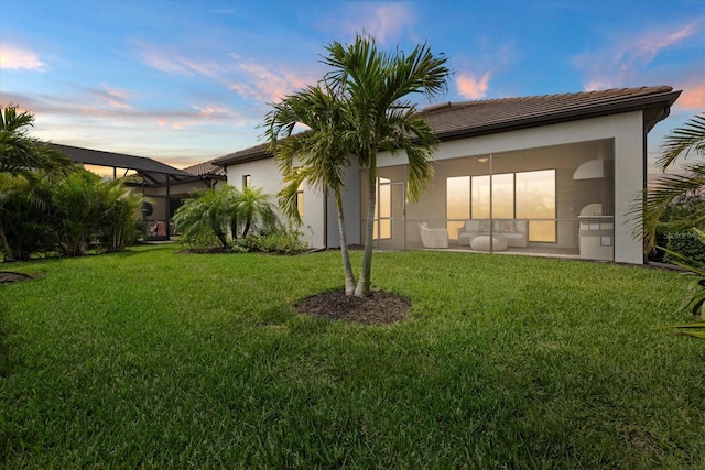 back of property at dusk with a lawn, a tiled roof, a lanai, and stucco siding