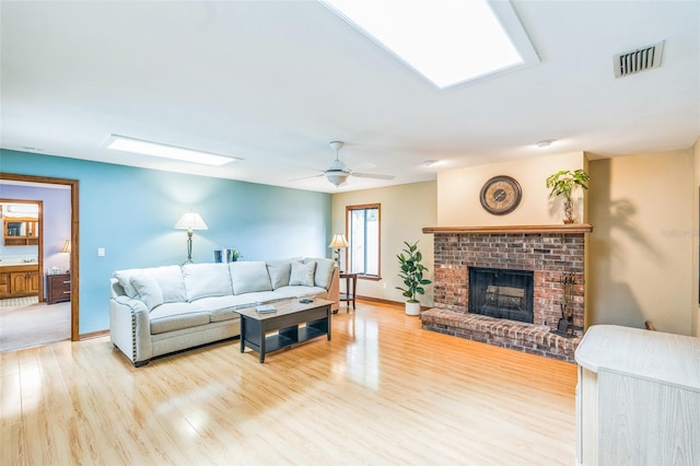living room with a skylight, light wood-type flooring, visible vents, a brick fireplace, and ceiling fan