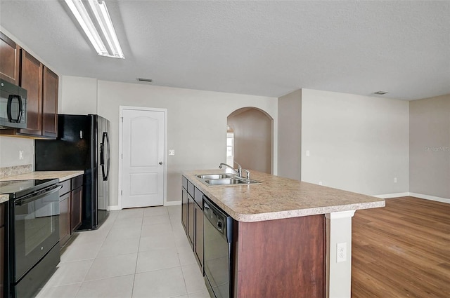kitchen with light countertops, a kitchen island with sink, arched walkways, a sink, and black appliances