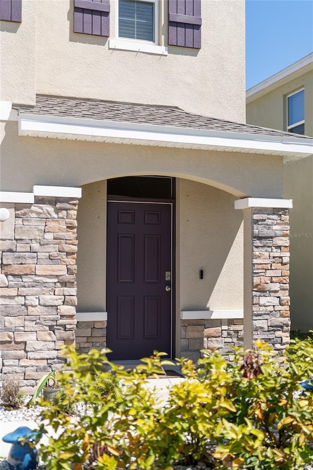 doorway to property featuring stone siding, a shingled roof, and stucco siding