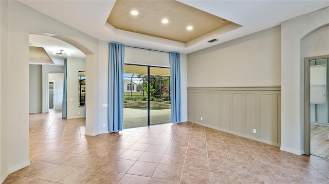 empty room featuring light tile patterned flooring, a wainscoted wall, a raised ceiling, visible vents, and arched walkways