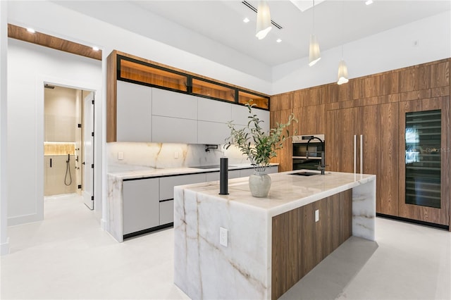 kitchen with decorative backsplash, modern cabinets, a kitchen island with sink, light stone counters, and hanging light fixtures