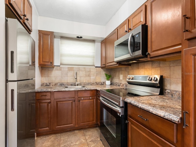kitchen featuring a sink, brown cabinetry, stainless steel appliances, decorative backsplash, and light stone counters
