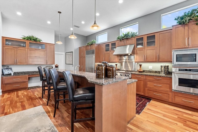 kitchen with built in appliances, a towering ceiling, under cabinet range hood, light wood-style floors, and brown cabinetry
