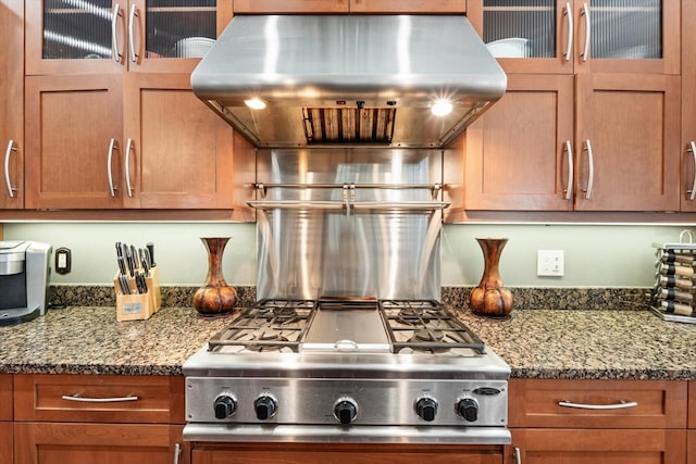 kitchen with dark stone counters, glass insert cabinets, brown cabinetry, ventilation hood, and stainless steel gas cooktop