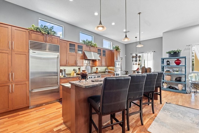 kitchen with built in appliances, a breakfast bar, under cabinet range hood, brown cabinetry, and light wood finished floors