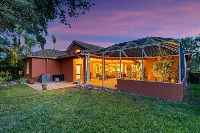 back of house at dusk featuring a patio area, a tiled roof, a lawn, glass enclosure, and stucco siding