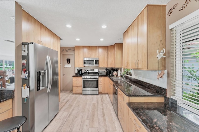 kitchen featuring backsplash, light brown cabinetry, appliances with stainless steel finishes, light wood finished floors, and dark stone counters