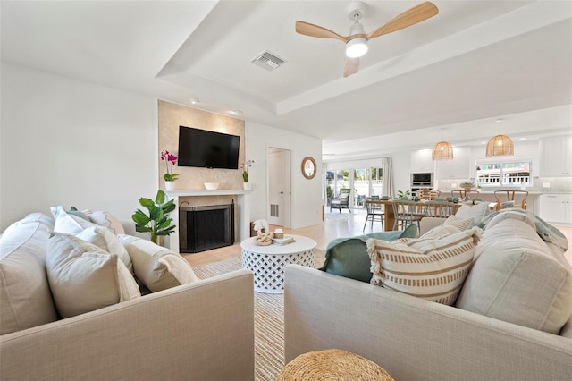 living area featuring a large fireplace, a ceiling fan, light wood-style floors, a tray ceiling, and visible vents