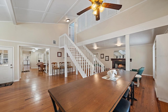 dining area featuring a warm lit fireplace, ceiling fan, a towering ceiling, stairs, and wood-type flooring