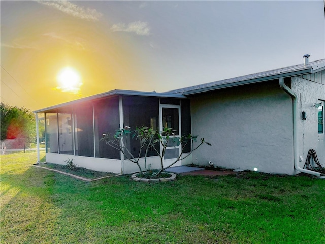back of house at dusk featuring a sunroom, stucco siding, fence, and a lawn