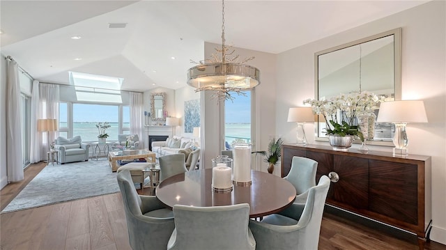 dining space with wood finished floors, a glass covered fireplace, a water view, an inviting chandelier, and lofted ceiling