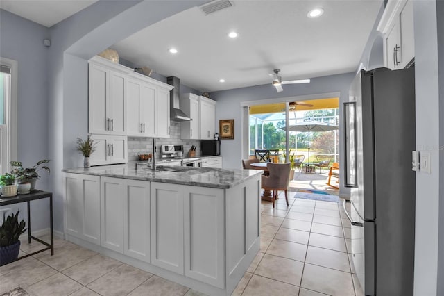 kitchen featuring freestanding refrigerator, wall chimney range hood, a ceiling fan, white cabinets, and light stone countertops