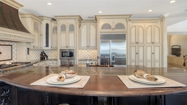 kitchen with built in appliances, premium range hood, cream cabinets, a sink, and backsplash