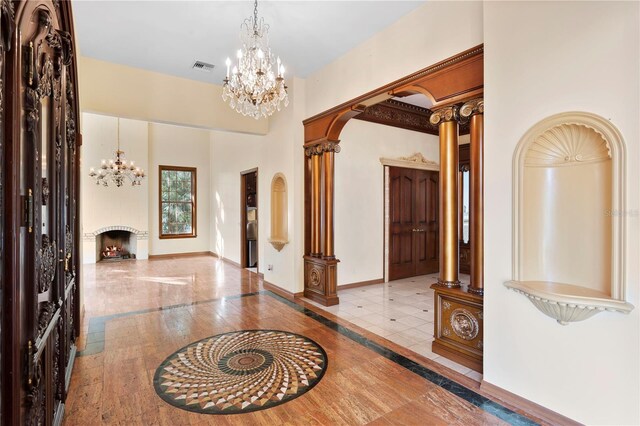 foyer featuring visible vents, baseboards, ornate columns, a notable chandelier, and a lit fireplace