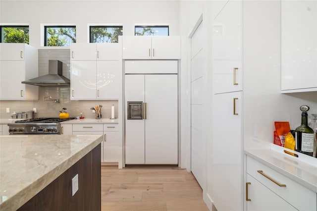 kitchen with white cabinets, wall chimney exhaust hood, paneled built in refrigerator, and stove