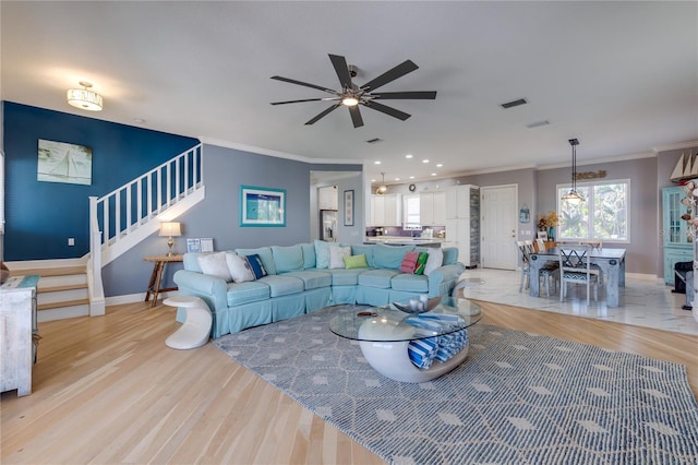 living area with stairway, a ceiling fan, light wood-type flooring, baseboards, and ornamental molding