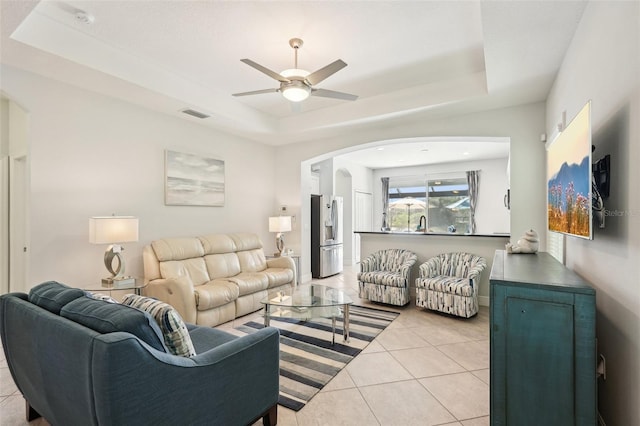 living room with arched walkways, light tile patterned floors, ceiling fan, visible vents, and a tray ceiling